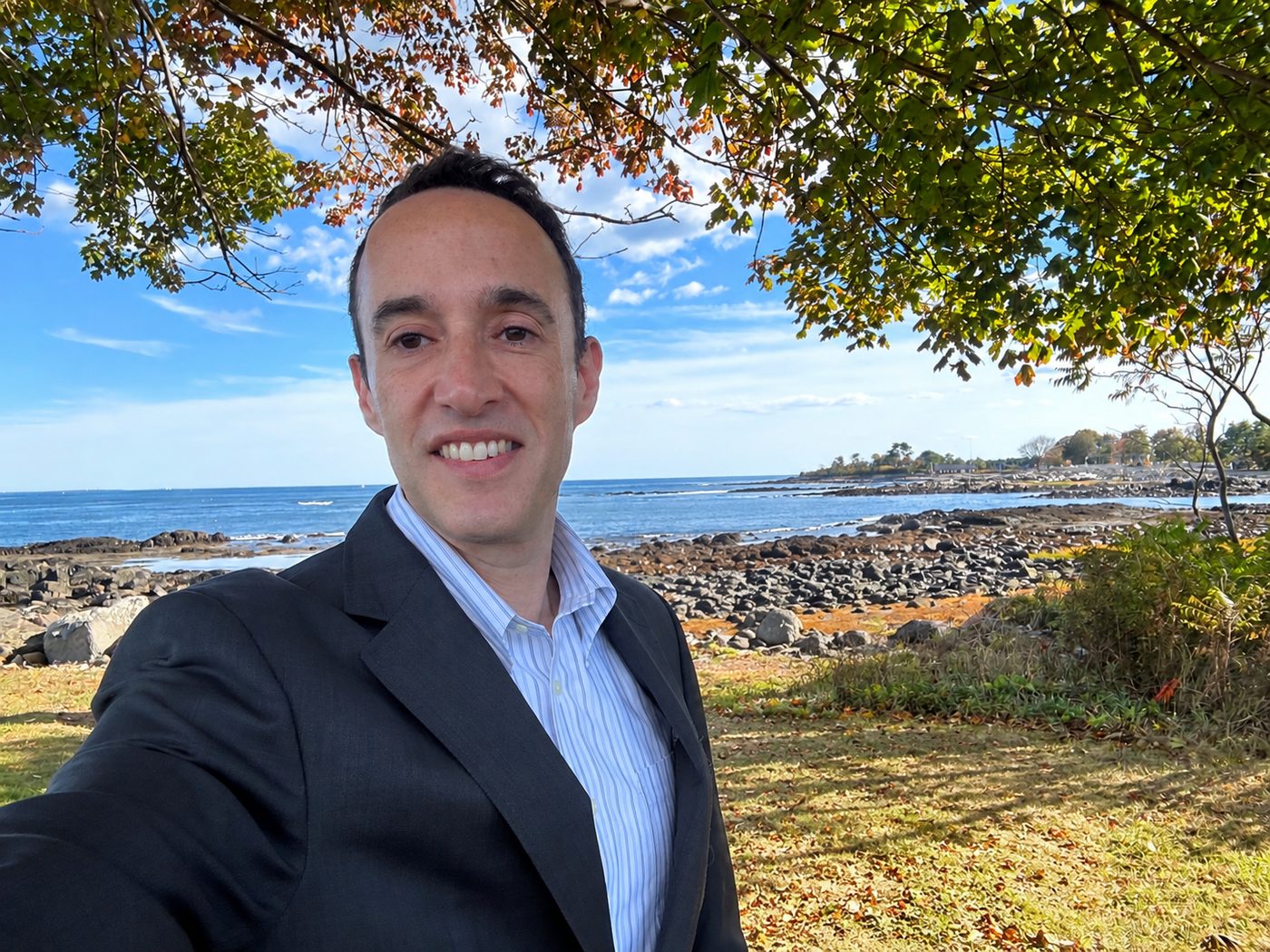 Andrew Kolidas smiling on a rocky New England shoreline on a bright autumn day